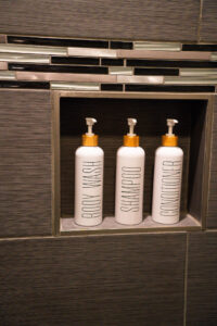 3 refilable white bottles with pump dispenser and the words Body Wash, Shampoo and Conditioner on them stood on a tile shelf in a shower enclosure the tiles are dark grey with a band of glass mosaic tiles above the shelf